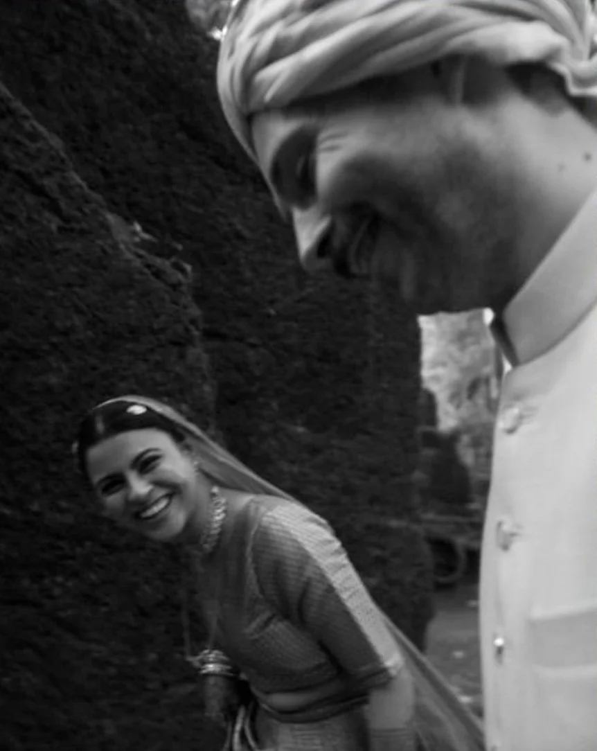 A bride in a dupatta smiles at the groom in a turban, black-and-white wedding photo